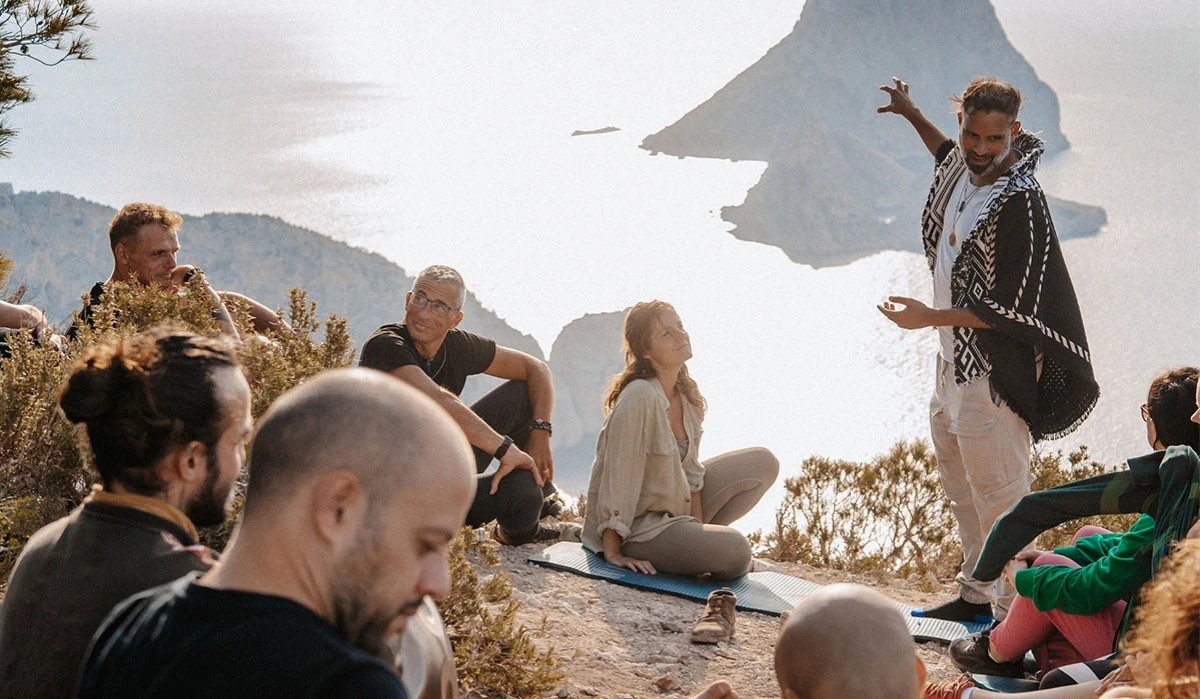 A SOMA Breath facilitator leading an outdoor group session on a clifftop above the sea, with participants seated on mats around him.