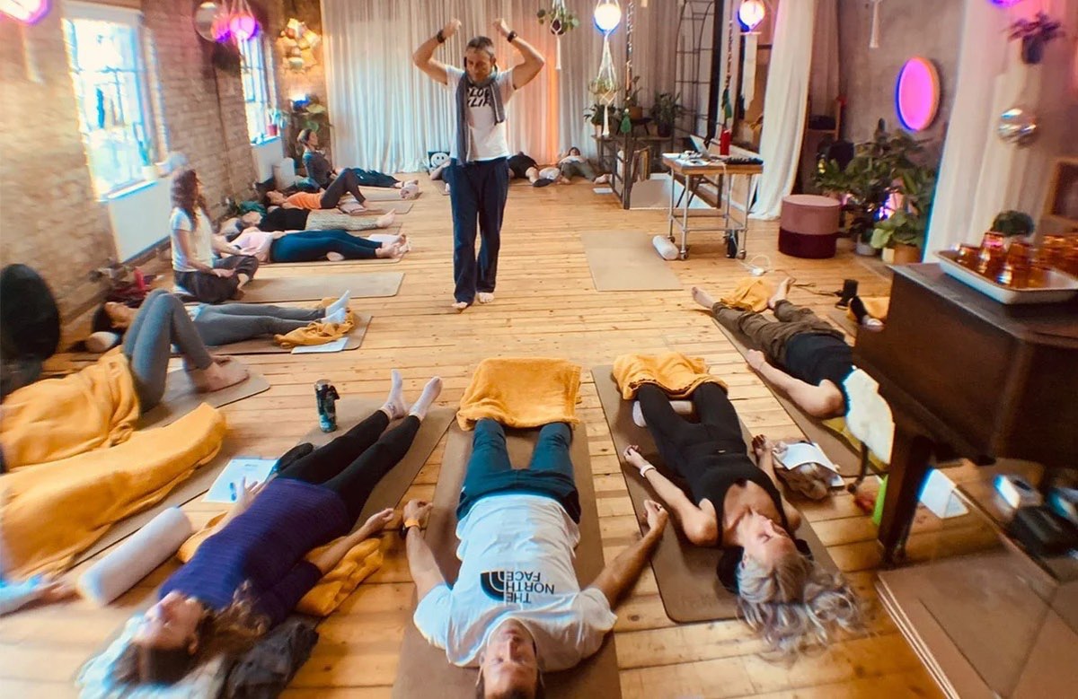 Participants lying on mats across a wooden studio floor during a SOMA Breath session, with a facilitator standing at the front of the room.